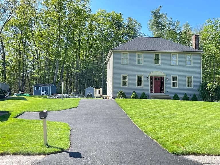 Exterior of 102 Maria Ave Southbridge MA — two-story colonial with light gray-blue siding, red front door, and mature wooded backdrop